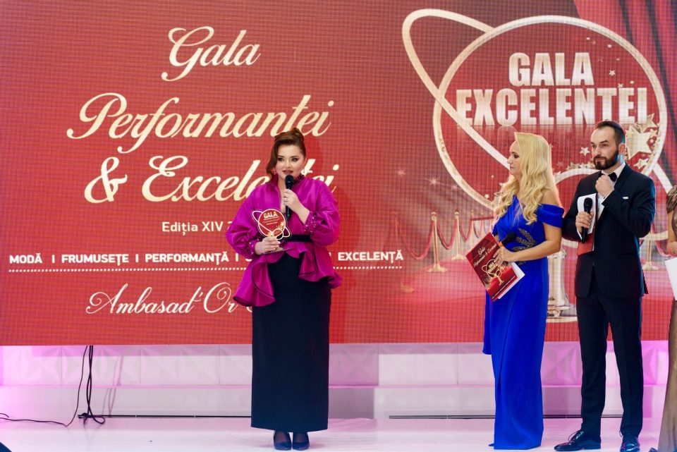 Three people stand on the stage of the Gala of Excellence and Performance 2024, with a red and gold backdrop displaying the event name. One person, a talented wedding dress designer, proudly holds an award.