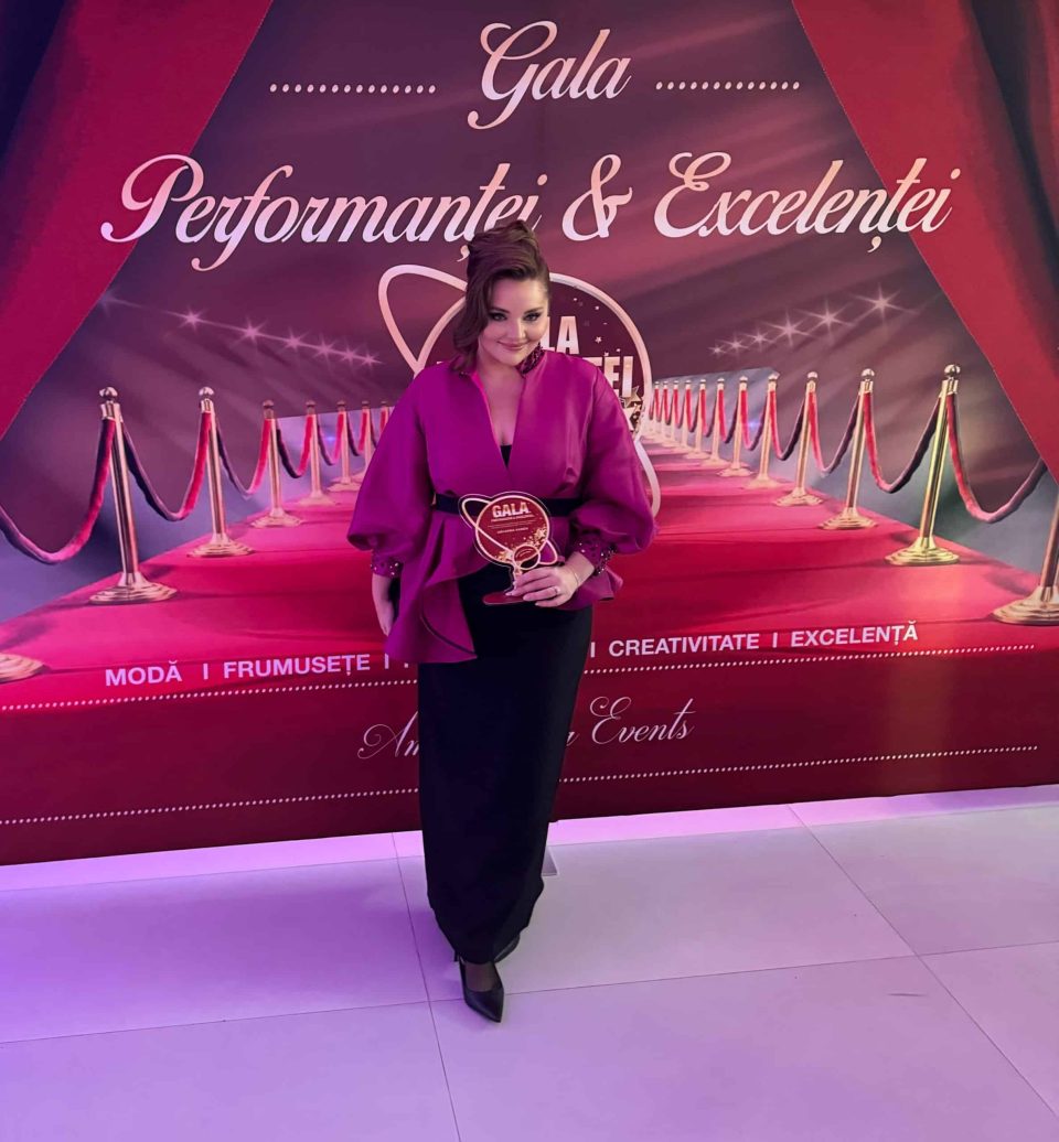 A person in a purple outfit poses in front of a red and violet backdrop 'Gala of Excellence and Performance 2024', holding a round award with pride, highlighting elegance synonymous with Romania.