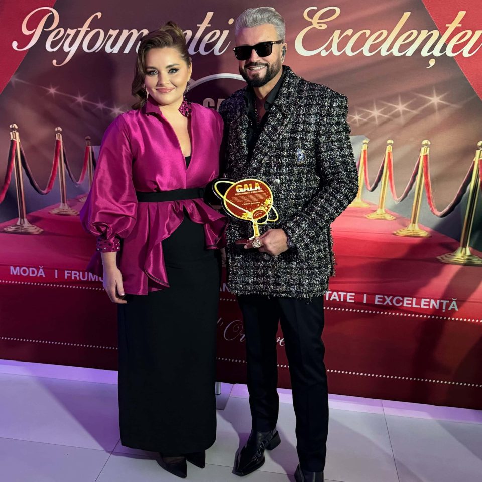 Two people posing; one wears a pink top and a black skirt, the other a textured jacket, holding a trophy. Against the red and gold backdrop with 'Performances and Excellences', they shine at the 2024 Gala of Excellence in Romania.