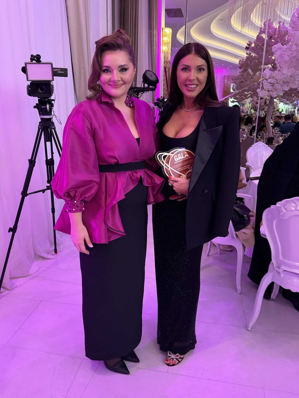 At Romania's Gala of Excellence and Performance 2024, two women pose. One shines in a shimmering magenta blouse, while the other stuns in a black dress with a blazer. Tables and decorative lights set the scene for this elegant indoor event.